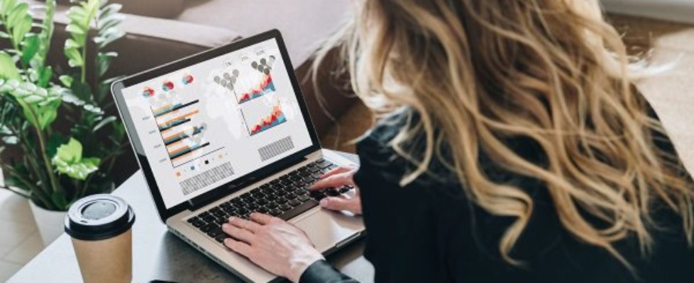 Rear view. Young businesswoman is sitting at table, working on laptop with graphs, charts, diagrams, schedules on screen